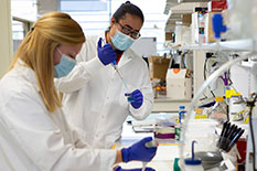 Two masked lab workers wear white lab coats and wear blue gloves while working with pipets