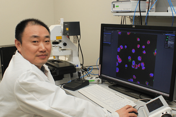 Dr. Shawn He sits at a computer next to a microscope. The screen displays what he sees on the microscope slide.