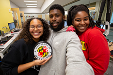 Three students smile while standing in a Terrapins Work 3d printing lab holding a 3d printed UMD seal
