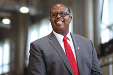 Dean Graham smiles while wearing glasses, a pinstripe gray suit jacket, a white collared shirt, and a red tie with white polka dots