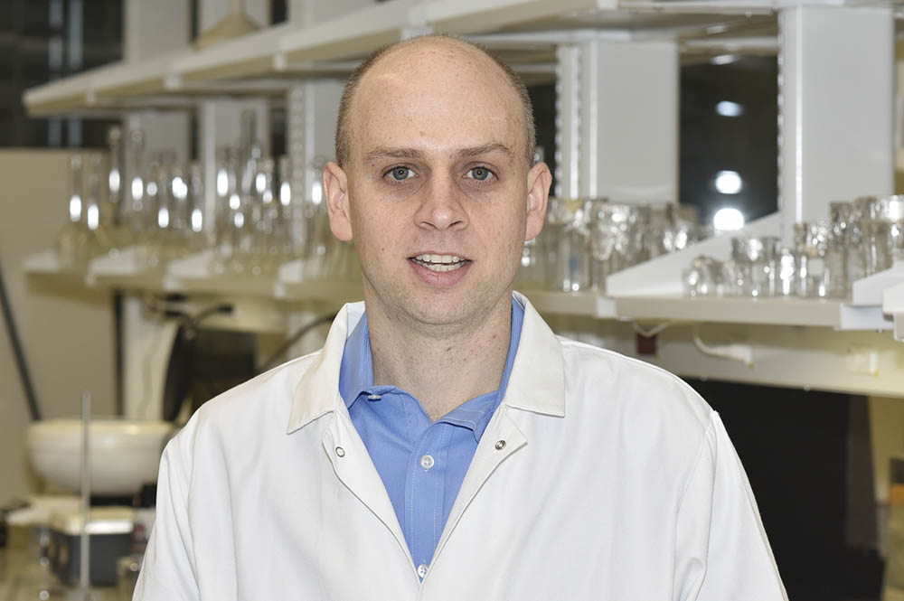 Dr. Chris Jewell is pictured in the lab wearing a white lab coat. Behind him are empty beakers and test tubes on shelves.