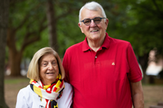 A woman wearing a patterned scarf in Maryland colors and a white shirt stands next to a man in a red polo. They are smiling at the camera.