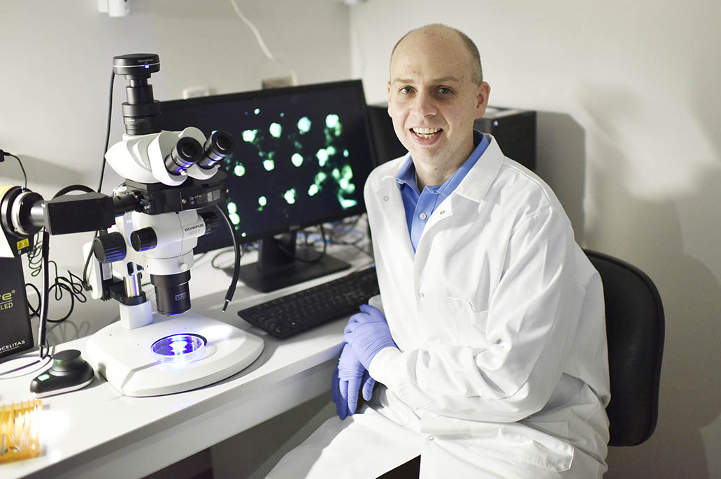 Dr. Chris Jewell is pictured sitting alongside a microscope and a screen displaying cells.