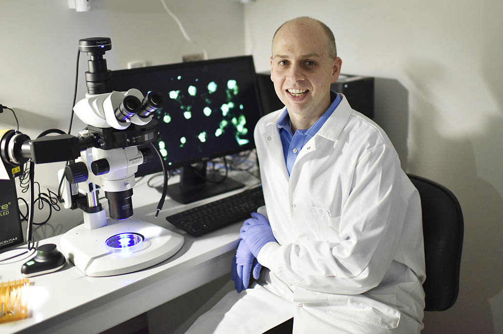 Dr. Christopher Jewell sits in a lab coat by a microscope and screen displaying cells.