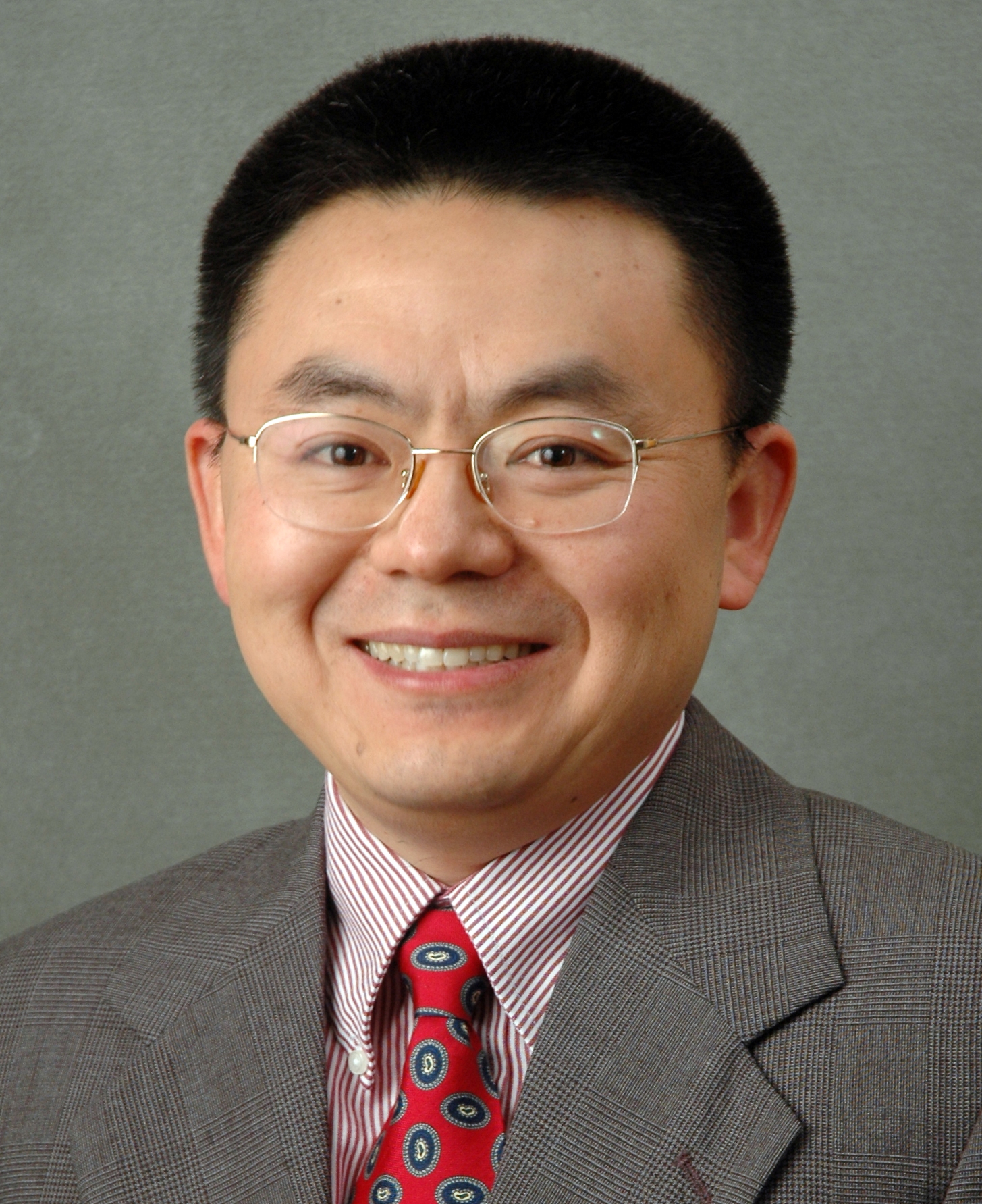 headshot of JC Zhao smiling and looking directly at the camera while wearing wire-rimmed glasses with a striped dress shirt, necktie, and dark suit jacket
