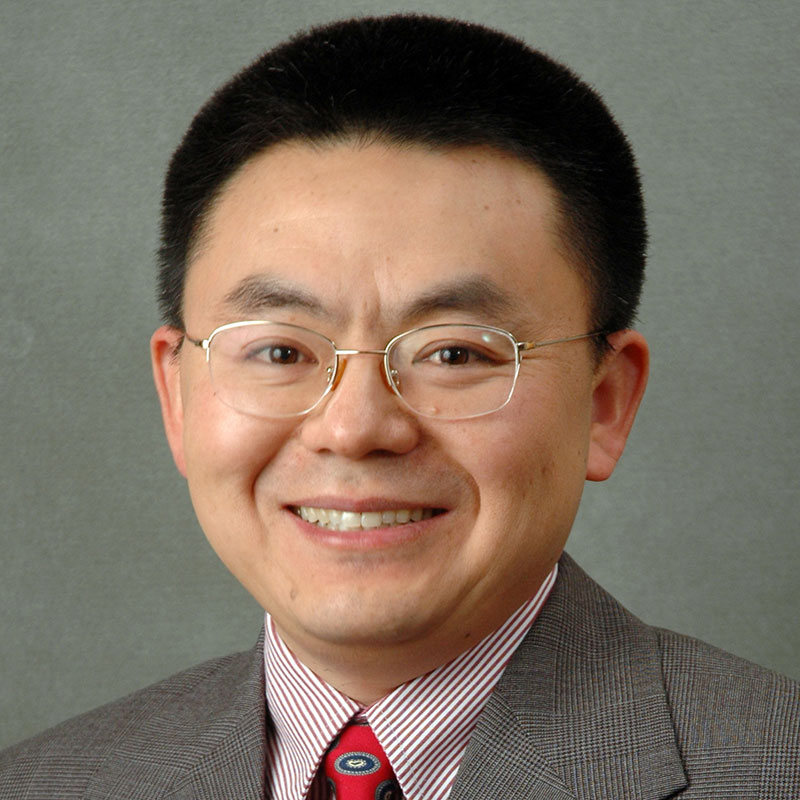 headshot of JC Zhao smiling and looking directly at the camera while wearing wire-rimmed glasses with a striped dress shirt, necktie, and dark suit jacket