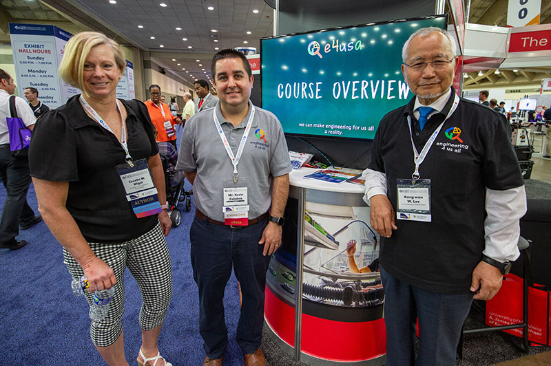 three members of the E4USA team wearing conference lanyards and standing at their booth in the exhibit hall at the 2023 ASEE Annual Conference