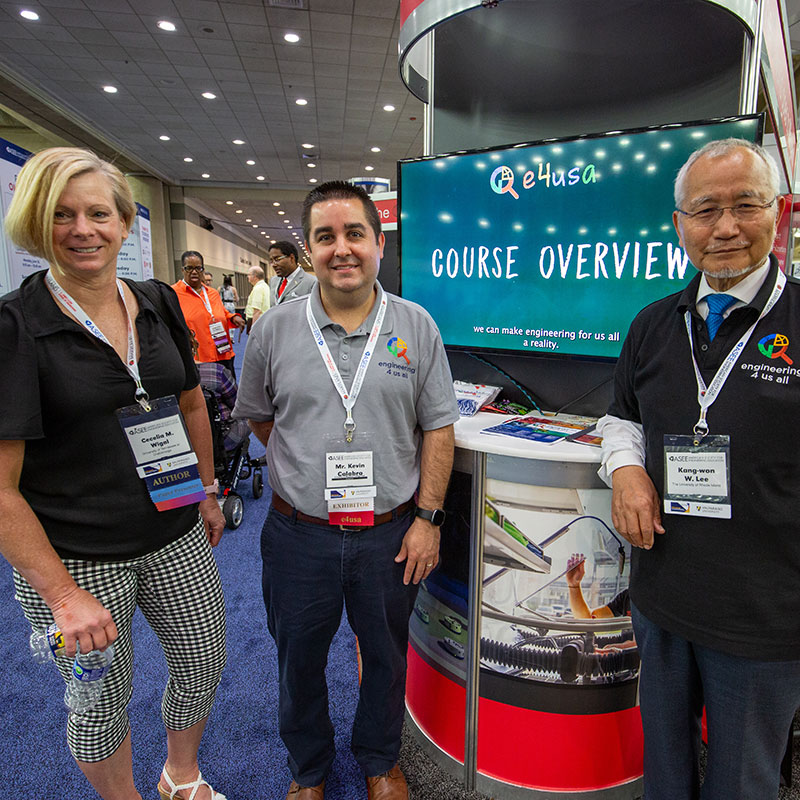 three members of the E4USA team wearing conference lanyards and standing at their booth in the exhibit hall at the 2023 ASEE Annual Conference