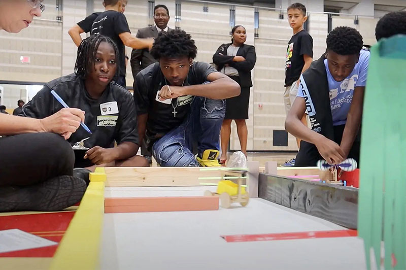 Rising ninth graders at Oxon Hill High School monitor the progress of their rubber band-powered cart in an engineering competition, while UMD president Darryll J. Pines observes in the background. Photo by Haimanot Yohannes Berhe.