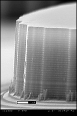 Christou’s laser mesas are constructed out of bands of different materials, each with a different function. At the base is a multilayered mirror that reflects all of the laser’s electromagnetic radiation up and out.  The laser itself is generated in the lower band, whose material is designed to have energy levels high enough to allow lasing—the stimulated emission of radiation, in this case infra-red—to take place. The upper layers of the mesa have been designed to allow all of the infrared light generated to be transmitted out, without filtering or diminishing. At the top of each mesa is an aperture approximately 10 microns wide, and above that, a 